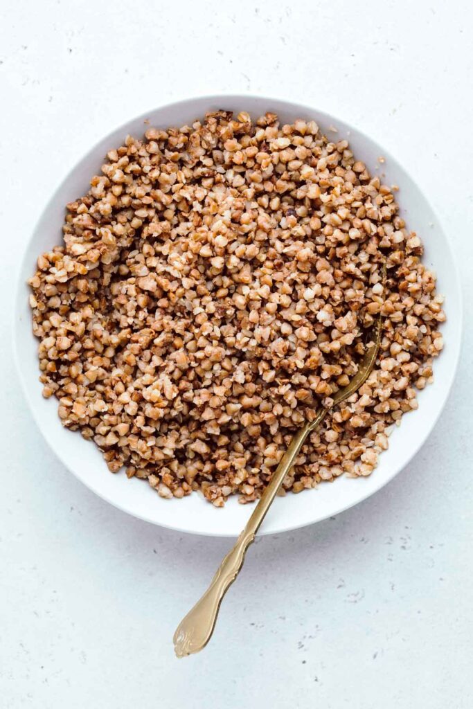 A large plate on a light grey backdrop with cooked roasted buckwheat in it and a gold colored fork in the buckwheat.