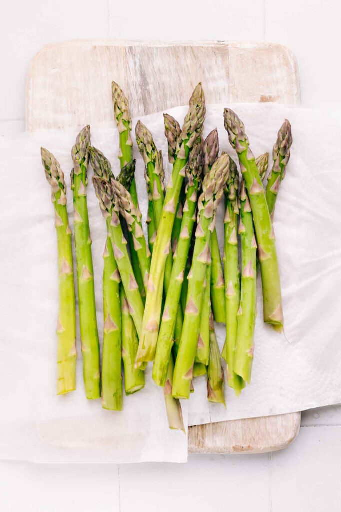 Asparagus on a white colored wooden cutting board placed on damp kitchen paper towels.