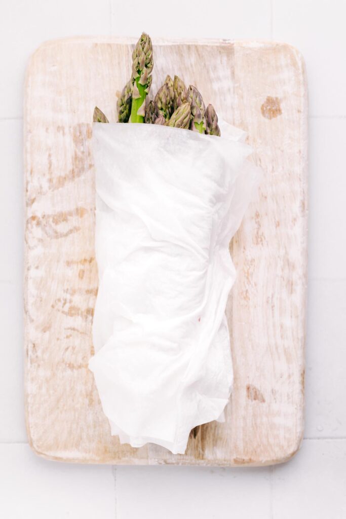 Asparagus on a white colored wooden cutting board bundled in damp kitchen paper towels.