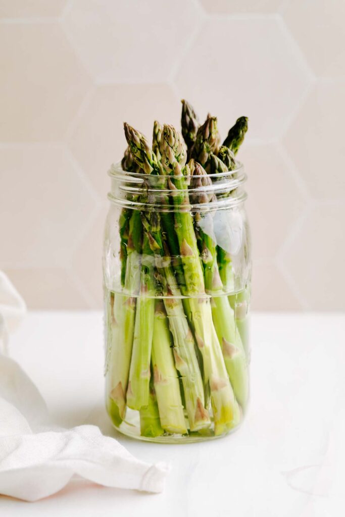 A mason jar filled with water with trimmed asparagus in it on a white backdrop.