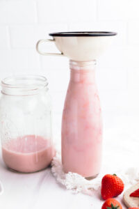 A large milk bottle with strawberry milk and a funnel on top next to a glass jar with a bit of strawberry milk on the bottom