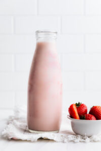 A milk bottle with pink strawberry milk on a white backdrop next to a small bowl with fresh strawberries