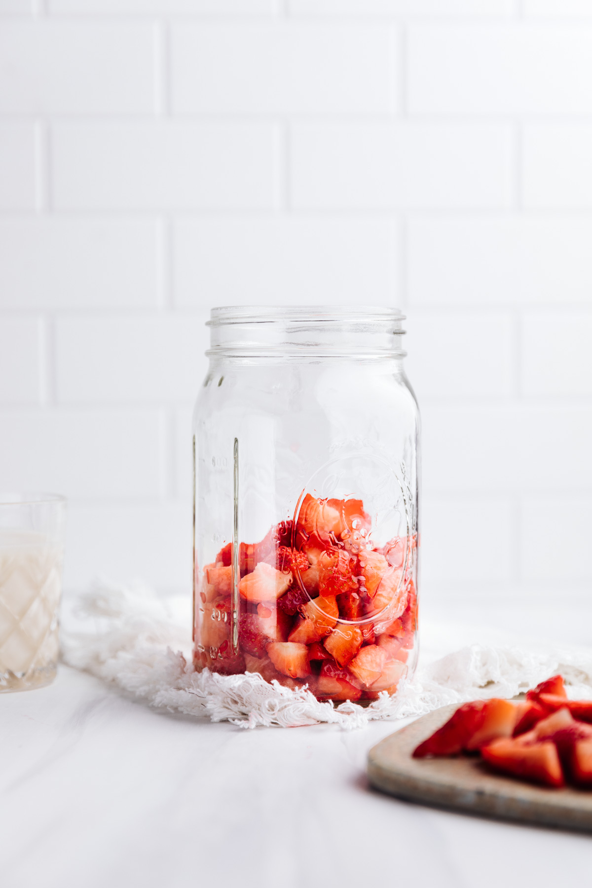A glass jar with chopped strawberries on the bottom on a white backdrop