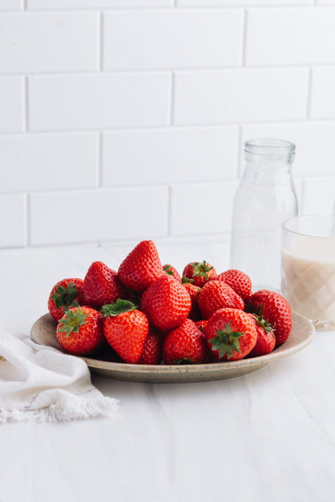A bowl with fresh strawberries on a white backdrop with a small glass of milk in the back