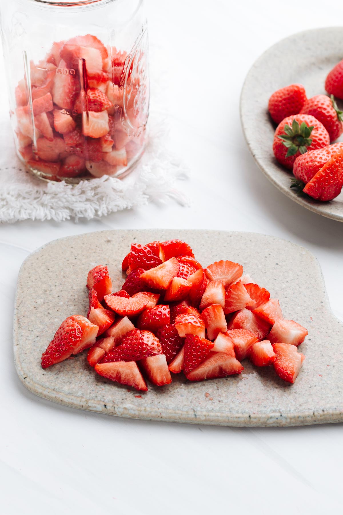 A small chopping board with chopped strawberries on a white backdrop with more fresh strawberry in the back