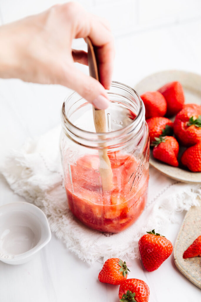 A large glass jar with chopped strawberries, sugar and a wooden spatula in the jar and a hand holding the top of the wooden spatula