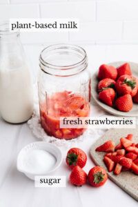 Ingredients for strawberry milk on a white backdrop in several small bottles, jars, a little small bowl, fresh strawberries and a small cutting board with chopped strawberries