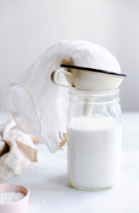 glass bottle with coconut milk and milk funnel with a nut milk bag with coconut milk pulp with white backdrop