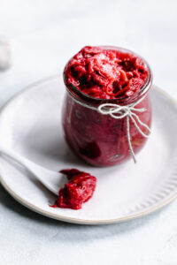 glass jar with red rhubarb jam on a white plate with a white teaspoon with rhubarb jam on it