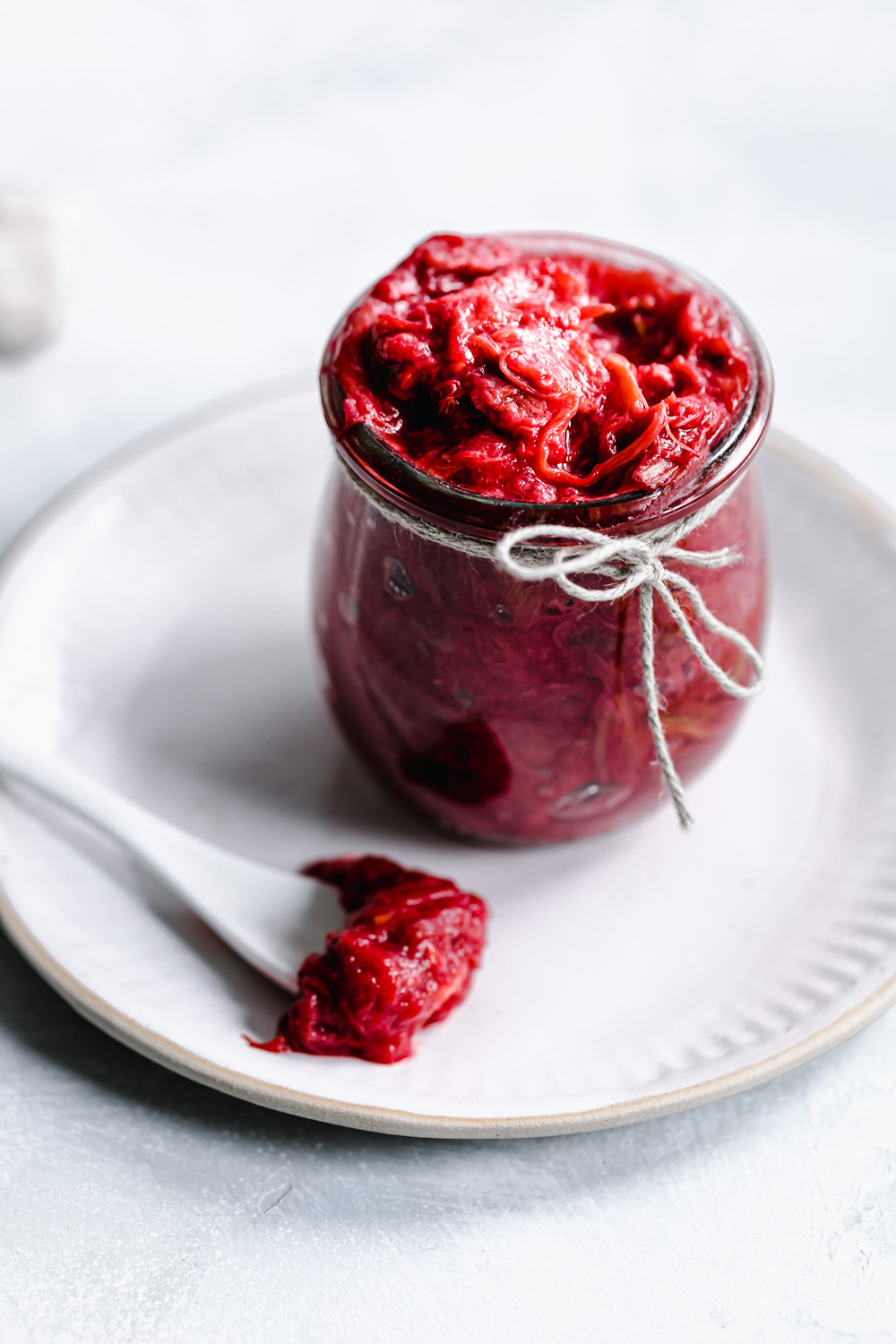 glass jar with red rhubarb jam on a white plate with a white teaspoon with rhubarb jam on it