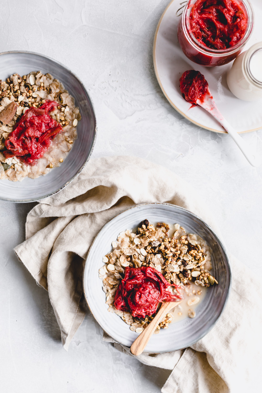 two blue bowls with granola and rhubarb jam on a white backdrop with rhubarb jam on a plate next to it