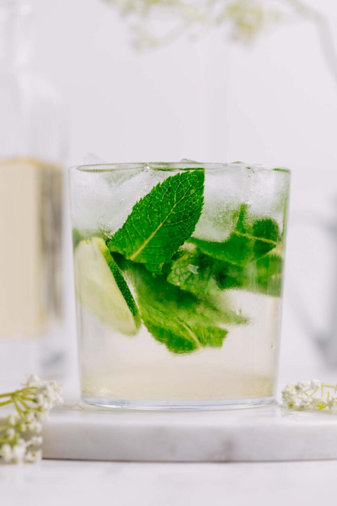 A glass with Hugo Spritz and with a lime wedge, mint leaves and ice cubes in the glass and elderflower decorations next to it.
