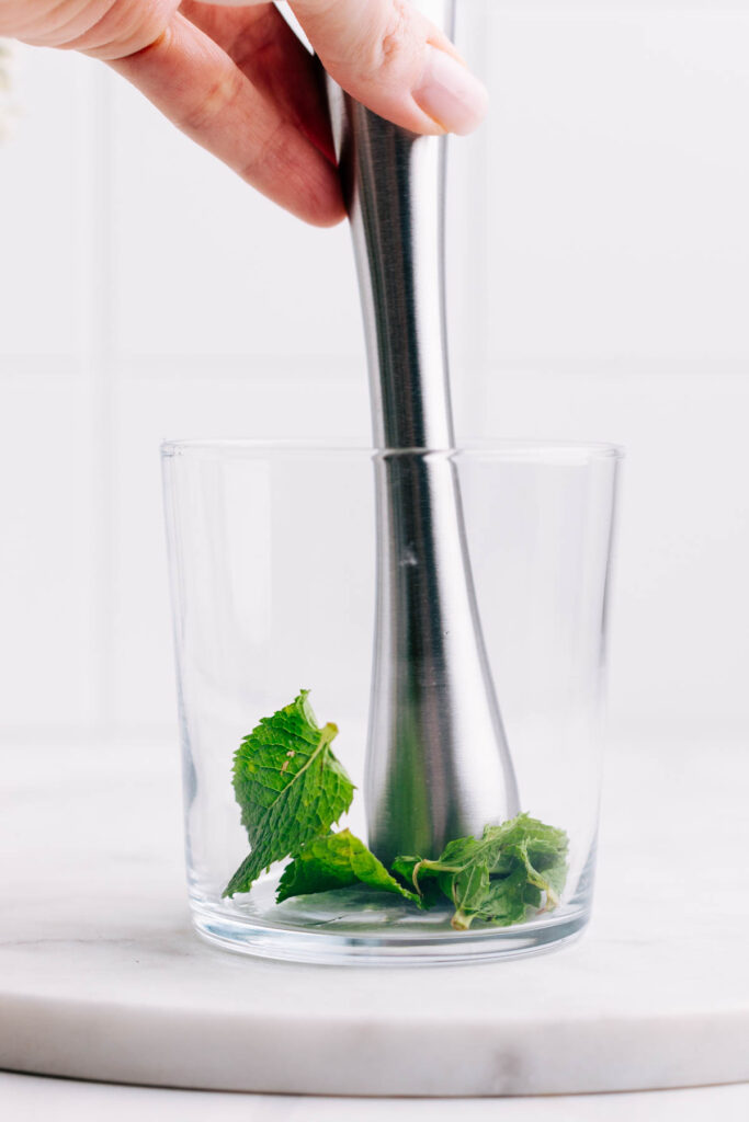 A glass with mint leaves and a cocktail muddler crushing tea leaves.