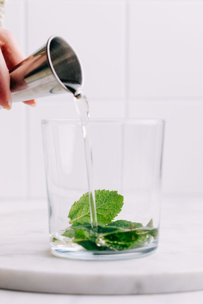 A glass with mint leaves and elderflower syrup being poured in top.