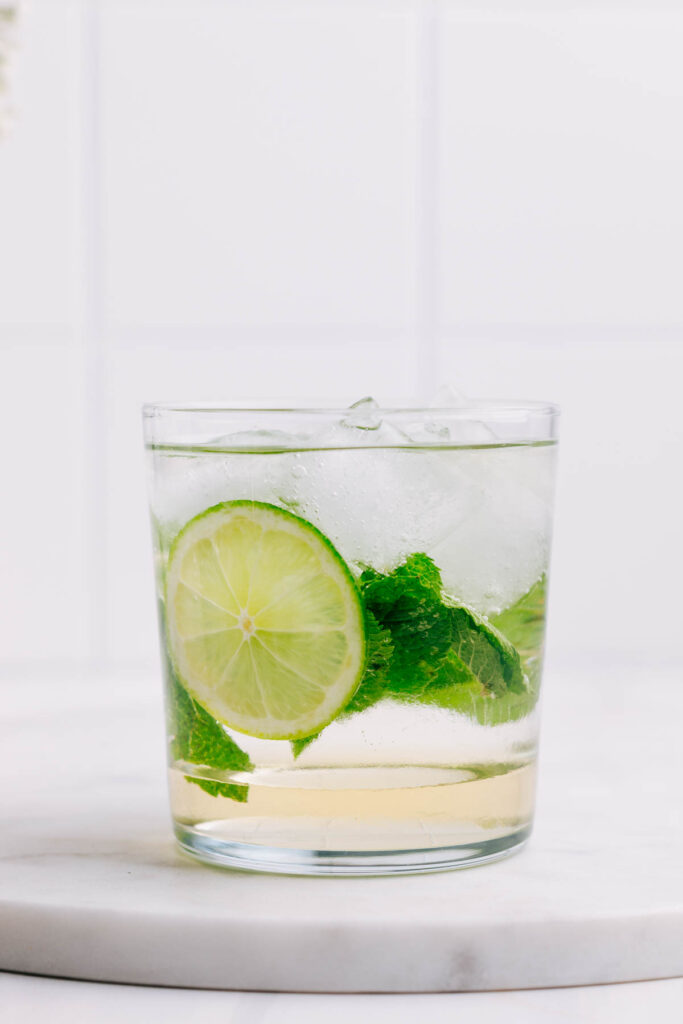 A glass with Hugo Spritz with visible layers of ice cubes, a lime wedge and mint leaves.