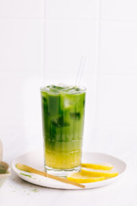 Matcha lemonade in a glass with ice cubes with a separate yellow and green layer with a glass straw on a white plate with lemon slices