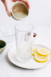 A glass on a white plate with lemon slices and a hand pouring in from a small bowl lemon juice in the glass