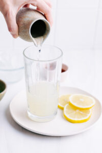A glass with a little bit of lemon juice on a white plate with lemon slices and a hand pouring in water in the glass