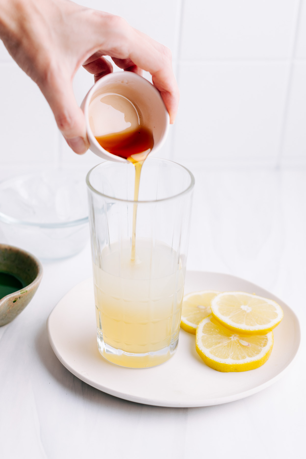 A glass filled half with lemon juice and water on a white plate with lemon slices and a hand pouring in maple syrup in the glass