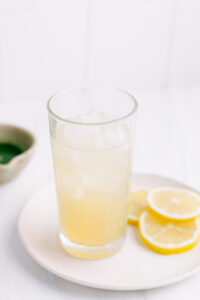 A glass with lemonade and ice cubes on a white plate with lemon slices on a white backdrop with a small bowl of matcha tea in the background
