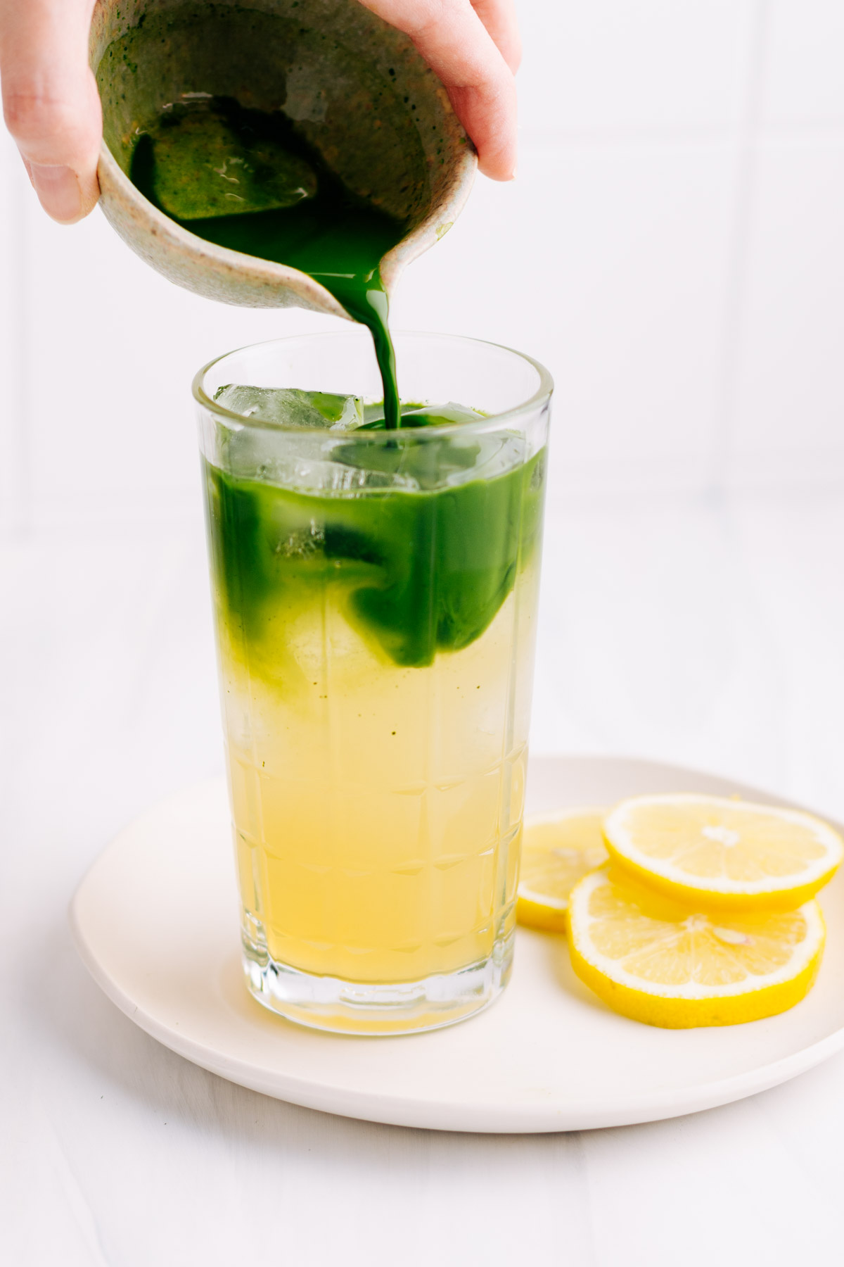 A glass with yellow lemonade and ice cubes on a white plate with lemon slices and a hand pouring in from a small bowl green matcha tea creating two separate colors in the glass