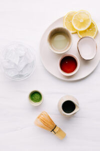 Ingredients for matcha lemonade on a white plate in several small bowls with lemon slices on a white backdrop with a bowl of ice cubes and a bamboo matcha whisk next to a small bowl with matcha powder