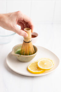 A small bowl with green matcha tea with a hand holding a matcha bamboo whisk in the bowl on a white plate with lemon slices