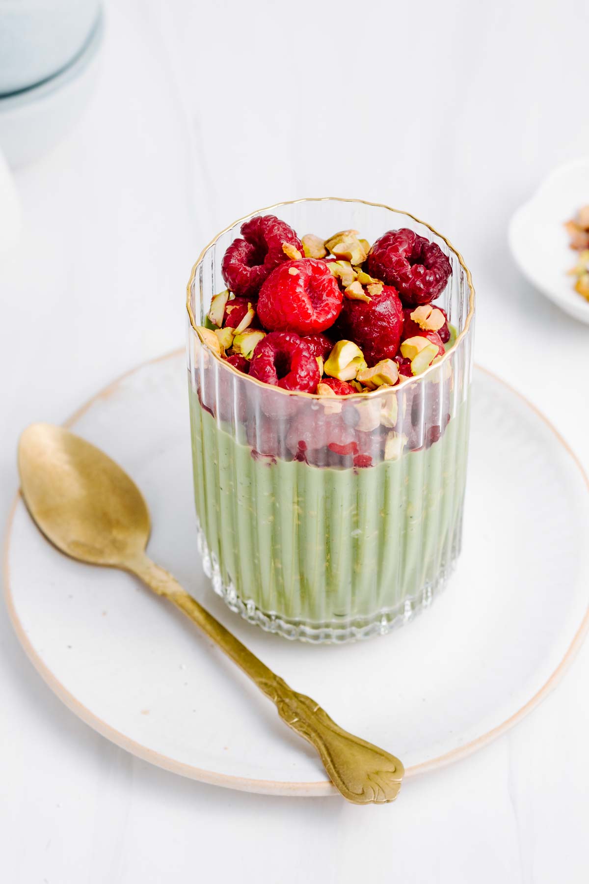 A glass filled with green matcha overnight oats and with raspberries and chopped pistachios on top on a white plate next to a golden spoon.