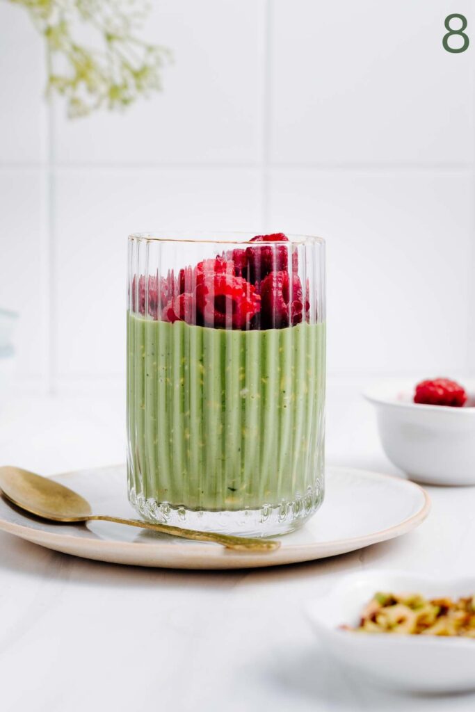 A glass with green matcha overnight oats and topped with raspberries on a white plate next to a golden spoon in front of a white backdrop.
