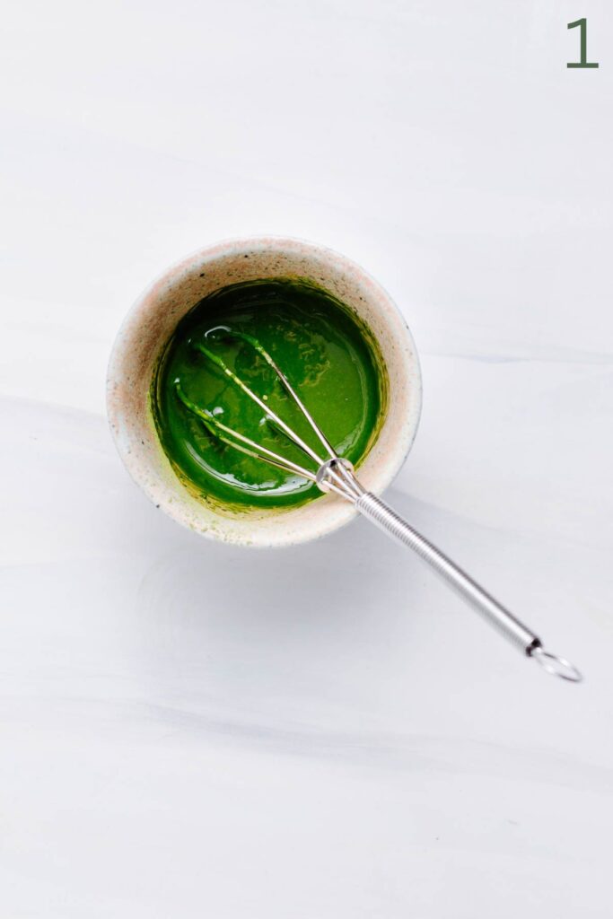 A small bowl with a green matcha mixture in it and a small stainless steel whisk on a white backdrop.