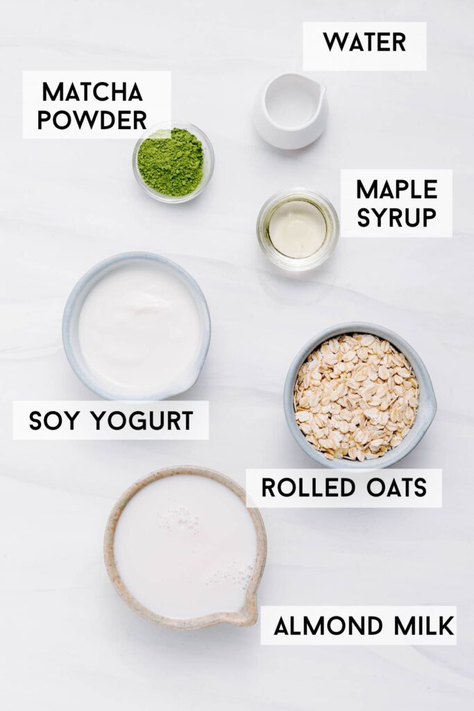 The ingredients for matcha overnight oats on a white backdrop in several bowls and the name of the ingredients in black bold written next to it.