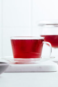 A glass teacup and plate on a white marble backdrop with red tea in it and a teapot with red tea in the background.