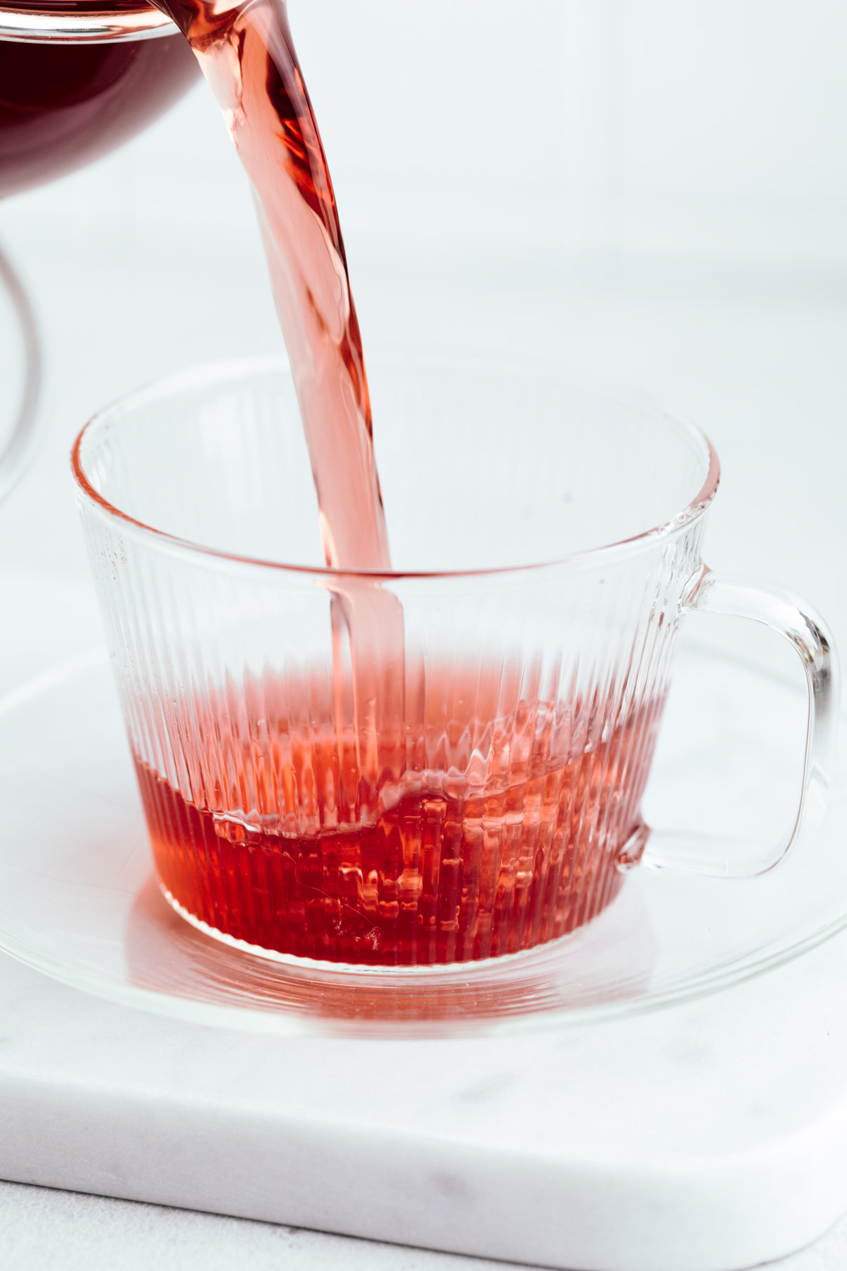 A glass teacup with a glass plate on a white marble backdrop with dark red tea being poured in the teacup.