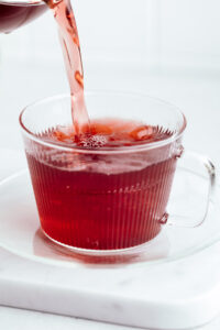A glass teacup with a glass plate on a white marble backdrop with dark red tea being poured in the teacup filling almost the entire teacup.