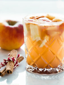 A glass filled with cider and apple chunks on a marble table with a cinnamon stick and an apple next to it.
