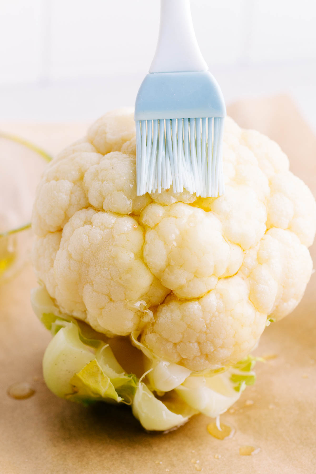Brown parchment paper with a whole head of cauliflower on it with a few drops of olive oil dripping on the bottom and a blue and white silicone pastry brush brushing olive oil on the cauliflower.