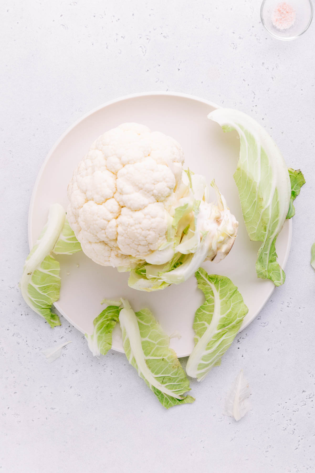 A head of cauliflower on a light grey backdrop on a round white plate with leaves of the cauliflower around it.