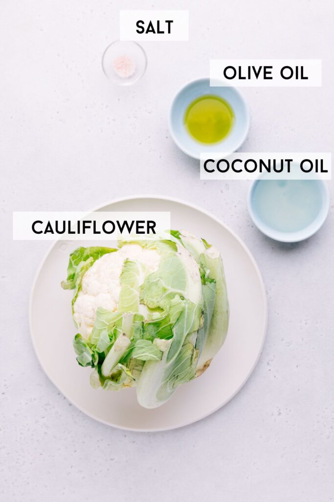 A light grey backdrop with a plate with a whole head of cauliflower and several small bowls with olive oil coconut oil and salt in it with the name of the ingredients next to it in bold.