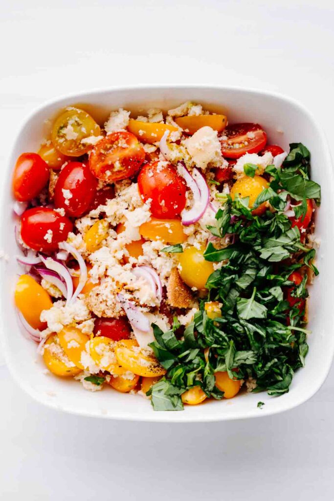 A square white bowl with crumbled wet bread, chopped yellow and red cherry tomatoes, sliced red onion and torn basil leaves.