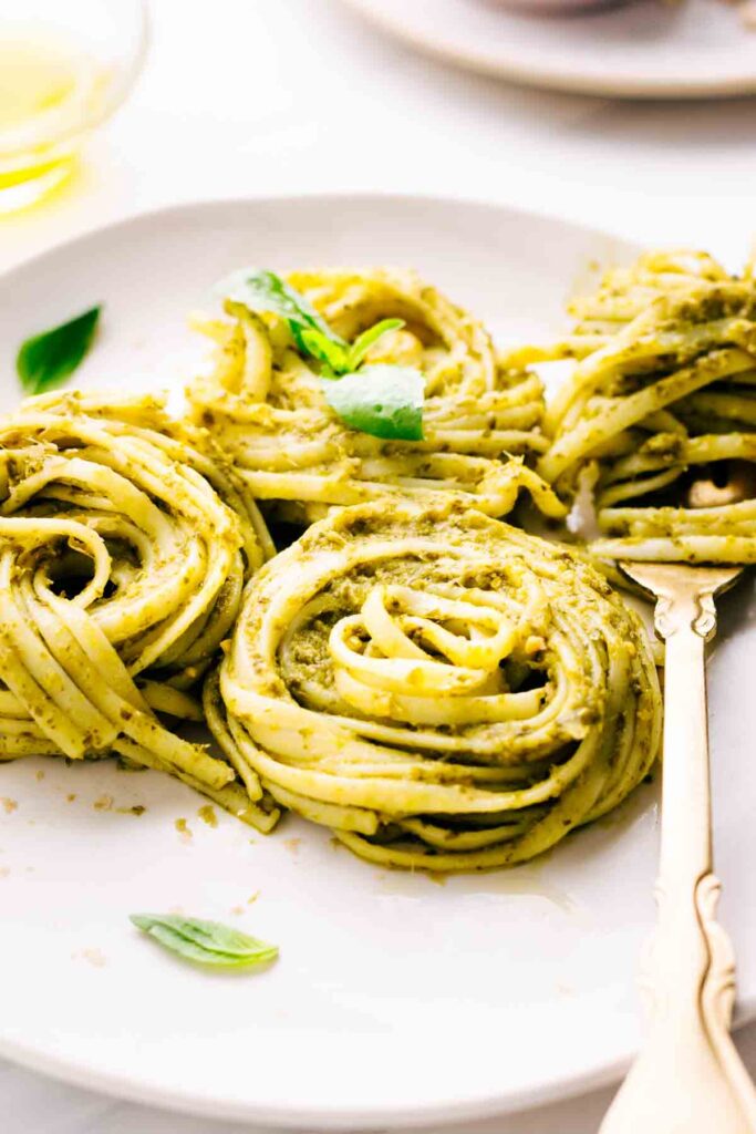 The close up of three swirls of pasta al pesto with basil leaves as a garnish next to a gold colored fork wrapped in more pasta al pesto.