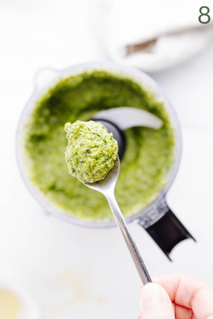 A spoon with pesto on it showing the texture of the pesto with a food processor with green basil pesto in the background.