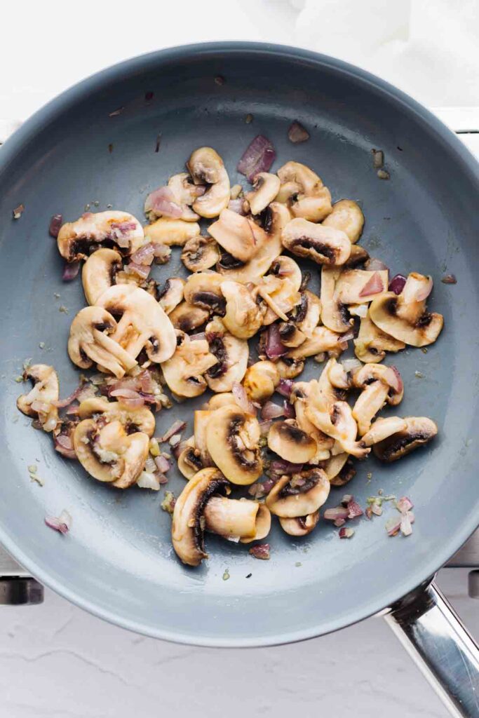 A blue pan with hot olive oil, diced red onion and browned button mushrooms.