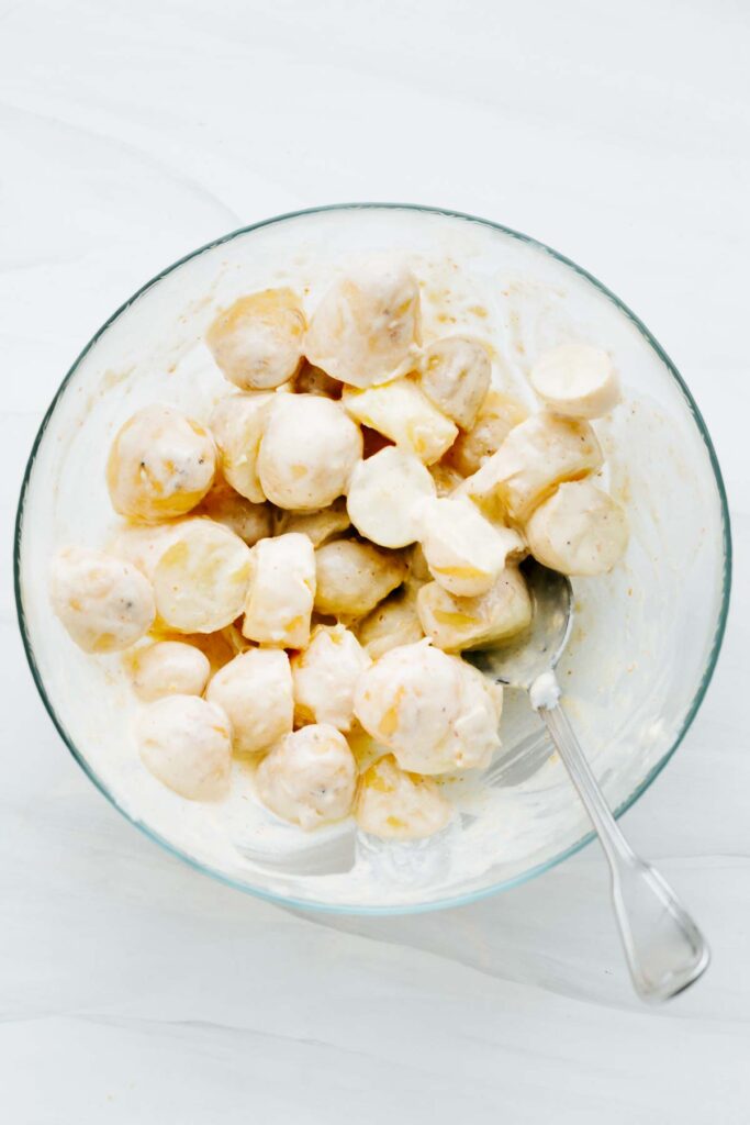 A glass bowl with a spoon and potatoes chunks mixed with a white dressing.