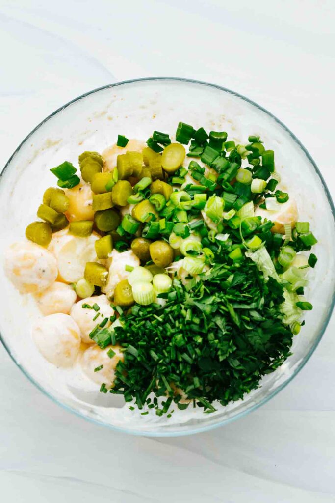 A glass bowl with potato chunks in a dressing topped with fresh herbs, cornichons, and chopped spring onions.