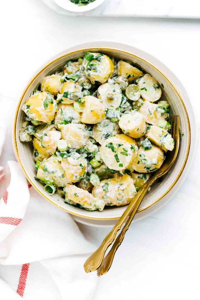 Potato salad with spring onions in a bowl with a gold fork and spoon on a white backdrop.