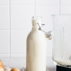 A milk bottle with potato milk next to a container from a blender with a few potatoes and a napkin and a white tile backdrop.
