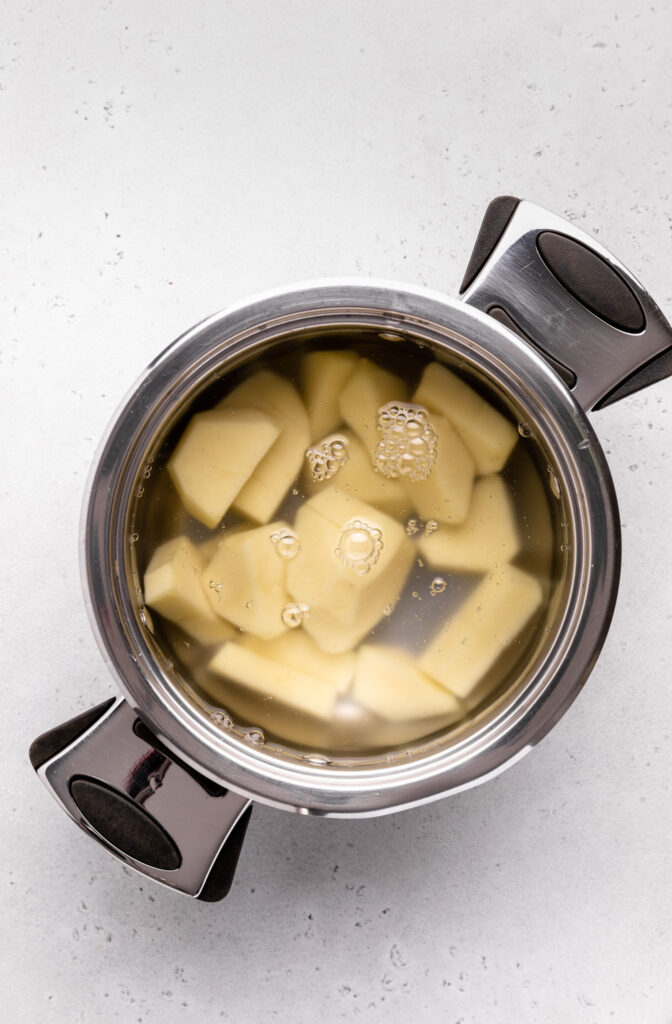 Potatoes in boiling water on a grey backdrop in a stainless steel pan.