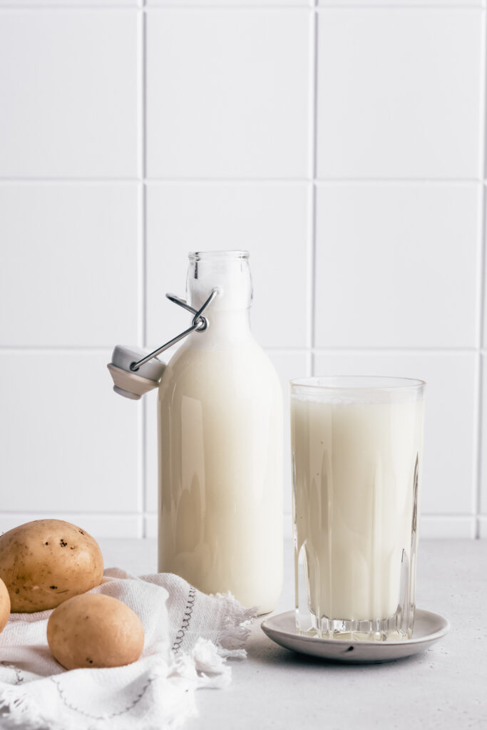 A milk bottle with potato milk next to a full glass with potato milk on a little plate with a few potatoes and a napkin and a white tile backdrop.