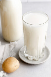 A glass with potato milk on a little plate on a light grey backdrop with a potato and a bottle of potato milk next to it.