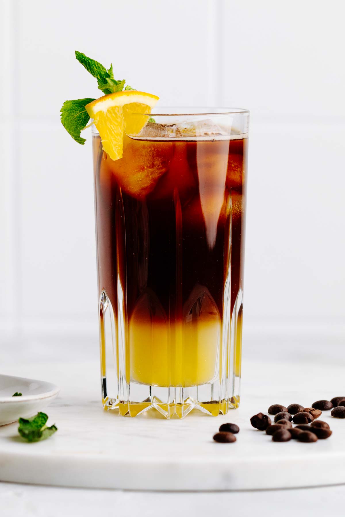 A tall glass with a two colored yellow and brown layered drink with a mint leave and orange wedge garnish on a white marble backdrop with coffee beans next to it.